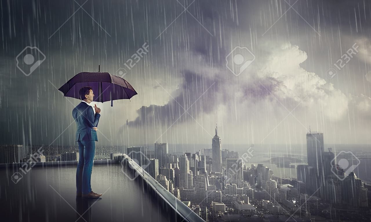 Nachdenklicher Geschäftsmann, der unter Sturm auf dem Dach eines Wolkenkratzers steht und den Horizont der Stadt beobachtet. Geschäftsforschungskonzept. Selbstbewusster Mann schützt vor Regen unter Regenschirm und blickt hoffnungsvoll nach vorne.