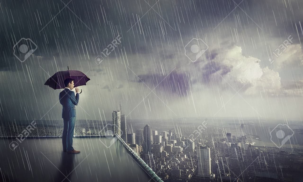 Uomo d'affari premuroso in piedi sotto la tempesta sul tetto di un grattacielo che guarda l'orizzonte della città. Concetto di ricerca aziendale. L'uomo fiducioso ripara dalla pioggia sotto l'ombrello, guardando fiducioso in avanti.