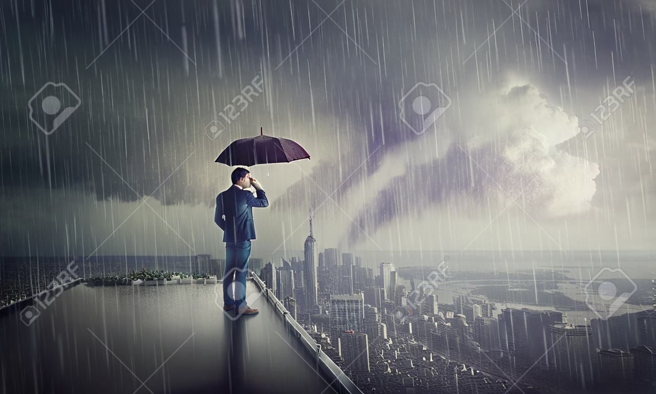 Nachdenklicher Geschäftsmann, der unter Sturm auf dem Dach eines Wolkenkratzers steht und den Horizont der Stadt beobachtet. Geschäftsforschungskonzept. Selbstbewusster Mann schützt vor Regen unter Regenschirm und blickt hoffnungsvoll nach vorne.