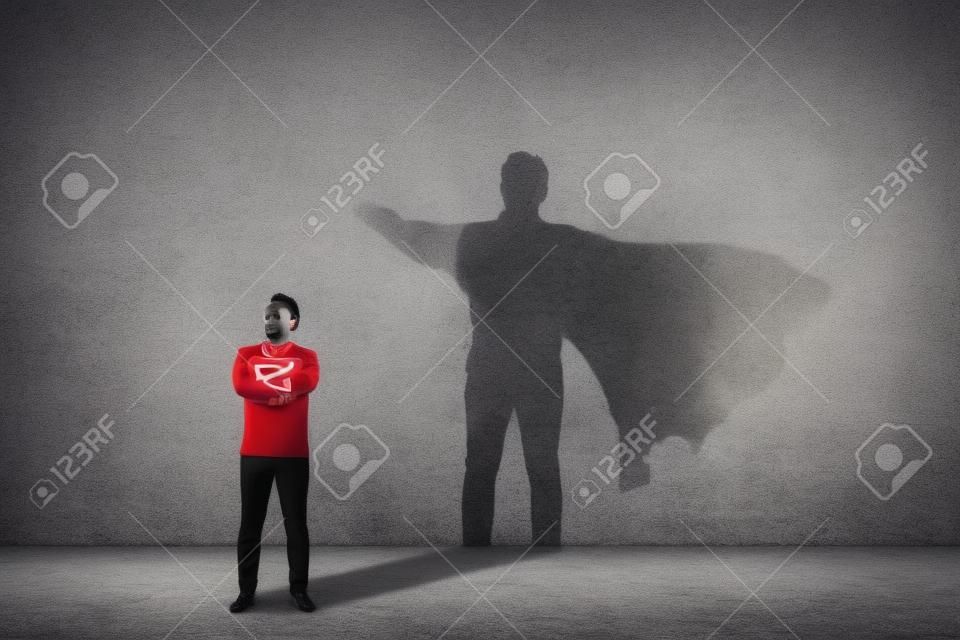 El hombre valiente mantiene los brazos cruzados, se ve confiado, lanzando un superhéroe con capa de sombra en la pared. Concepto de ambición y éxito empresarial. Poder de héroe de liderazgo, motivación y símbolo de fuerza interior.