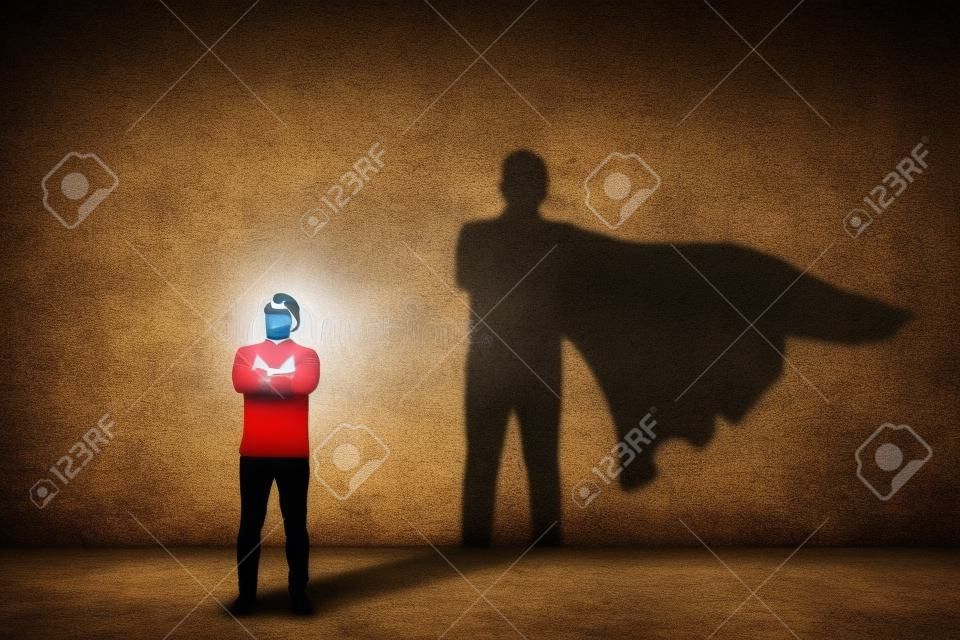 L'uomo coraggioso tiene le braccia incrociate, sembra sicuro di sé, proiettando un supereroe con l'ombra del mantello sul muro. Ambizione e concetto di successo aziendale. Simbolo di potere, motivazione e forza interiore dell'eroe di leadership.