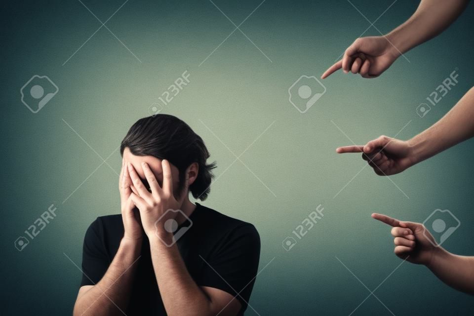 Disappointed and ashamed man, covers his face with palms, as many people fingers points to him from behind, blaming and scolding. Guy suffering emotional breakdown and depression, being under pressure
