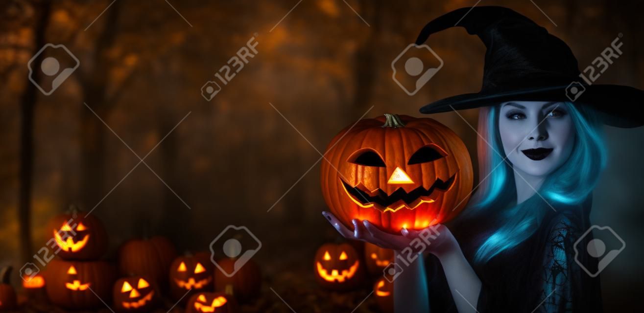 Halloweenowa czarownica z rzeźbiącymi dyniowymi i magicznymi światłami w ciemnym lesie