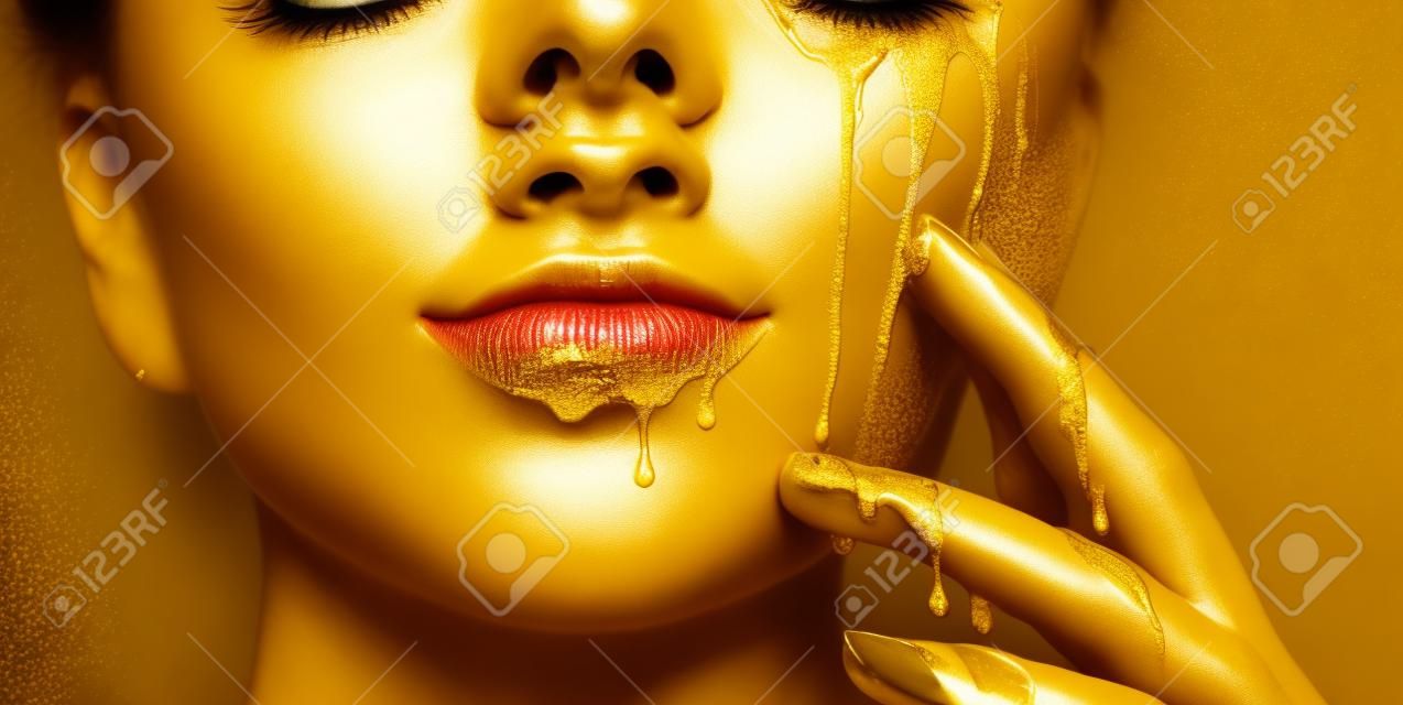 Goldene Farbflecken tropfen von den Gesichtslippen und der Hand, goldene Flüssigkeitstropfen auf dem Mund des schönen Modellmädchens, kreatives abstraktes Make-up. Schönheitsfrauengesicht