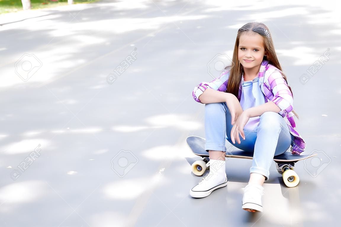 Smiling cute little girl child sitting with a skateboard. Preteen with penny board outdoors in summer day