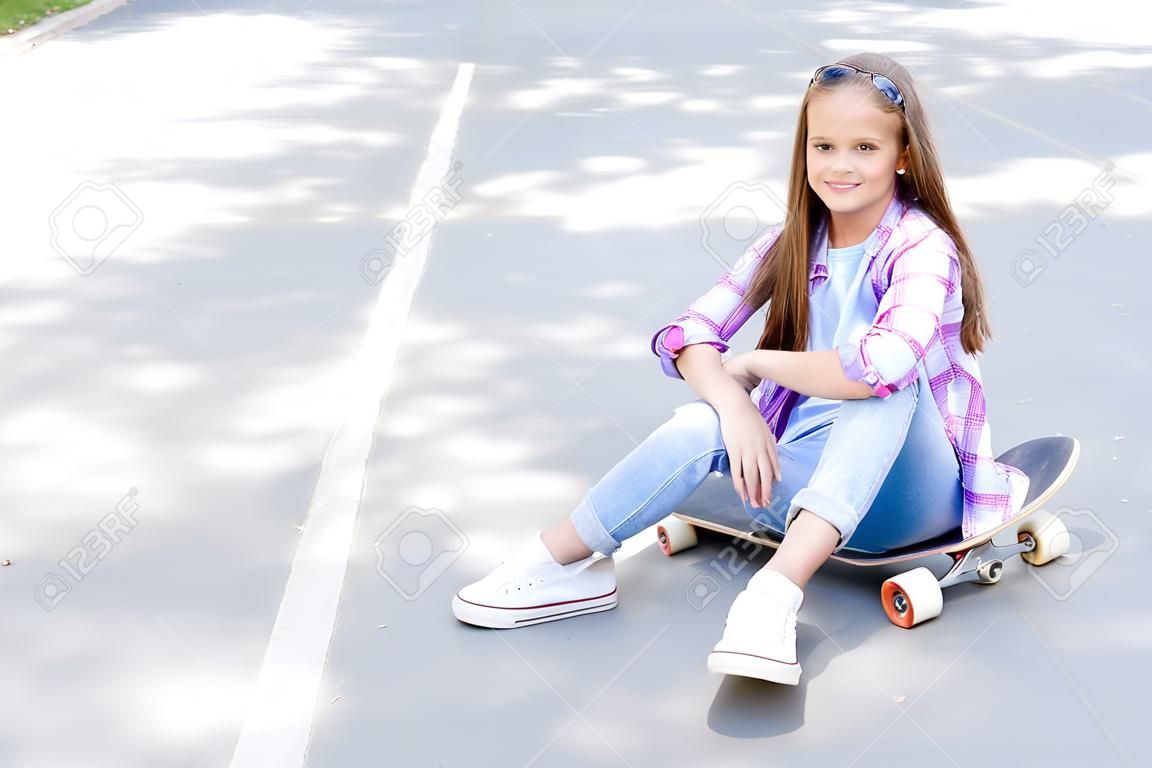 Smiling cute little girl child sitting with a skateboard. Preteen with penny board outdoors in summer day