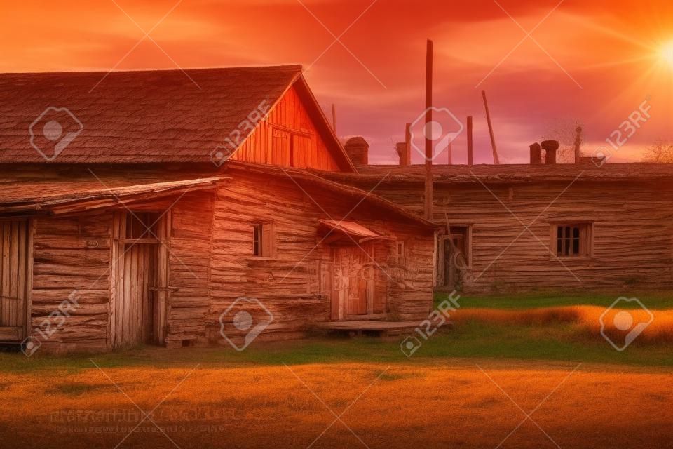 Courtyard of old wooden Russian house, barn with closed shutters in red warm light of sun, with shadows on dry grass. Abandoned village. Orange pink sunset