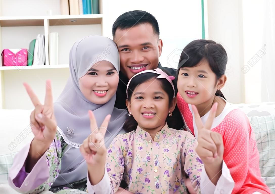 Happy Asian family at home. Muslim family showing v victory hand sign and having fun. Southeast Asian parents and children smiling.
