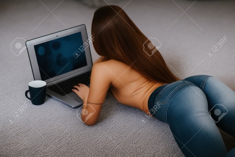 有完善的赃物的女孩在牛仔裤小短裤在家与在地毯的膝上型计算机一起使用。