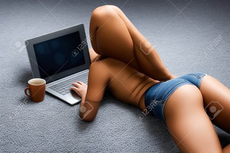 有完善的赃物的女孩在牛仔裤小短裤在家与在地毯的膝上型计算机一起使用。
