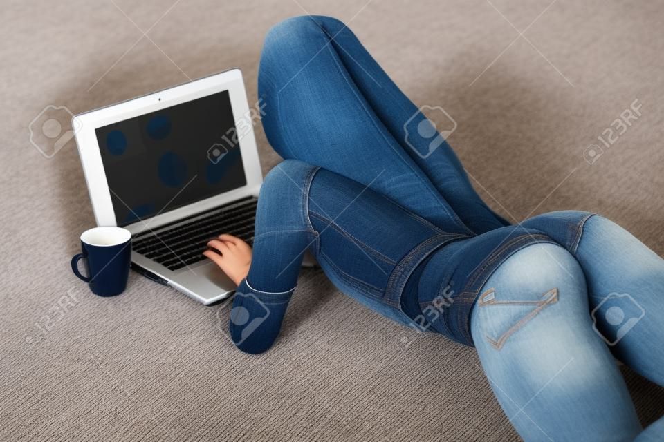 Chica con botín perfecto en jeans pantalones cortos trabajando con la computadora portátil en la alfombra en casa.
