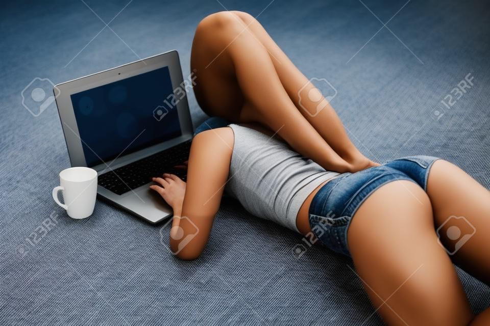 Ragazza con il bottino perfetto in pantaloncini di jeans piccoli che lavorano con il computer portatile sul tappeto a casa.