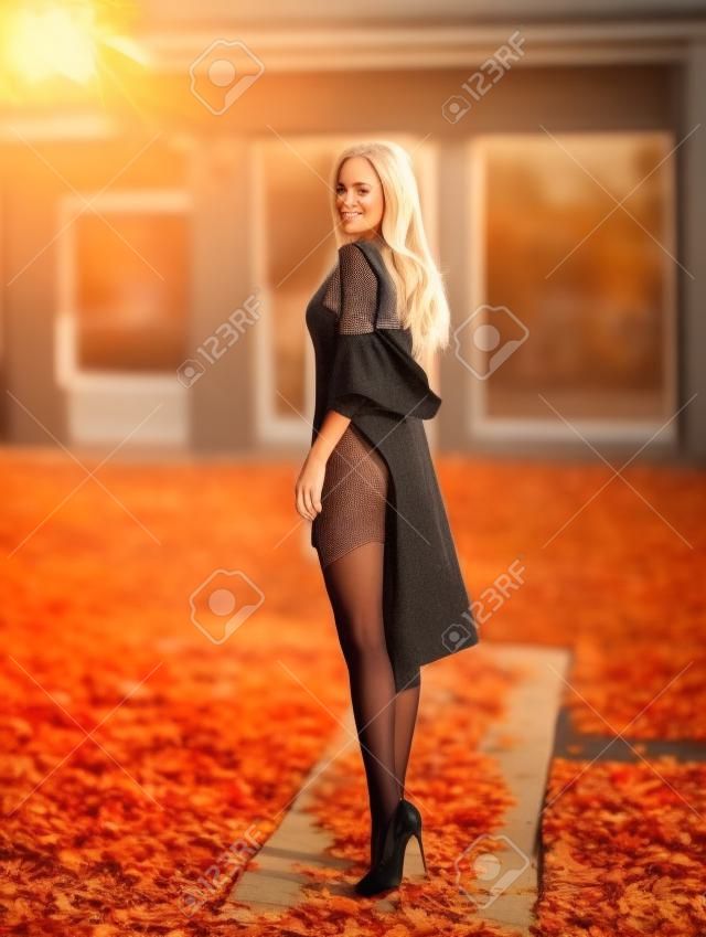 Mulher loira bonita com pernas perfeitas na meia-calça posando ao ar livre na rua de outono nas luzes do sol poente.