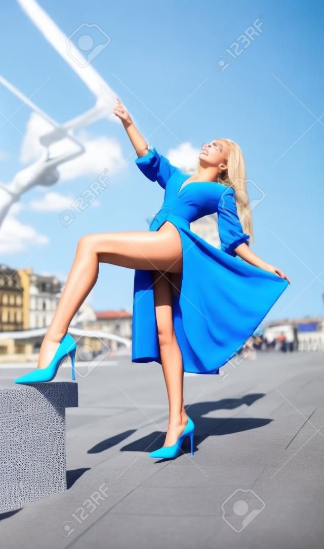 Beautiful blonde girl in the blue dress with perfect legs and shoes with high heels posing outdoor on the city square.