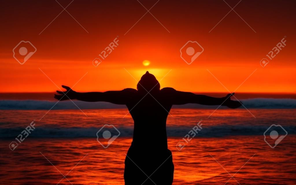A young man stands as a living shadow  in front of the ocean with his arms outstreatched to the orange sky and his head tilted back to absorb the setting sun 