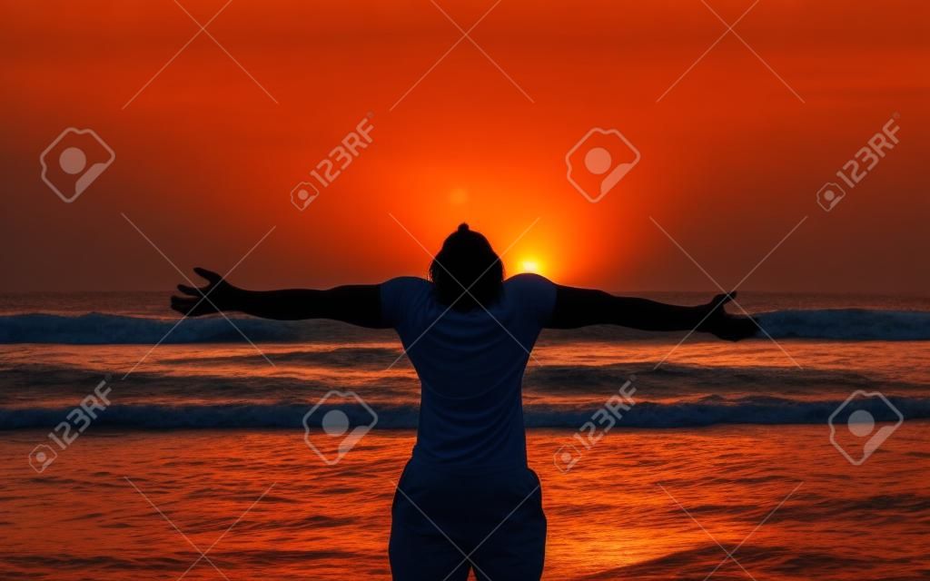 A young man stands as a living shadow  in front of the ocean with his arms outstreatched to the orange sky and his head tilted back to absorb the setting sun 