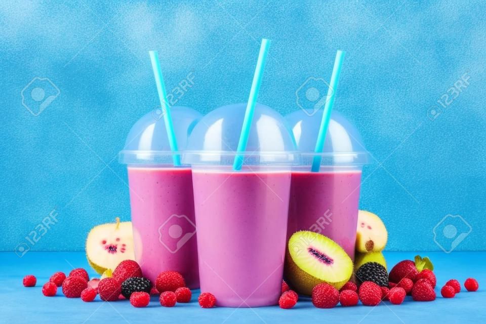 Fruit smoothies in plastic cups with blueberry, strawberry, kiwi, blackberry, raspberry and banana on blue background. Take away drinks concept.