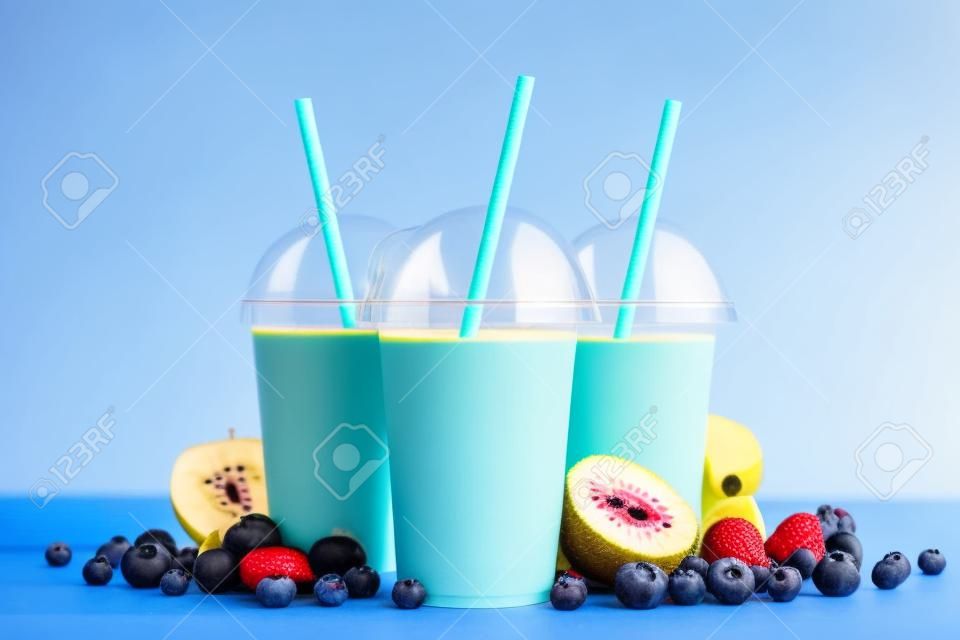 Fruit smoothies in plastic cups with blueberry, strawberry, kiwi, blackberry, raspberry and banana on blue background. Take away drinks concept.