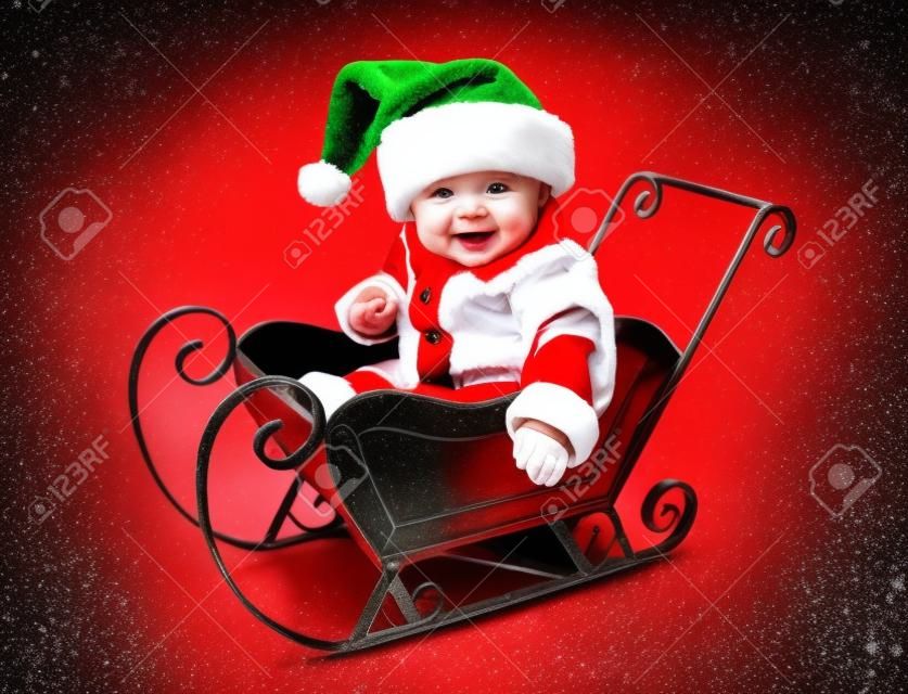 Adorable bebé joven vistiendo un traje de santa claus y sombrero, sentado en un trineo de nieve de Navidad metal