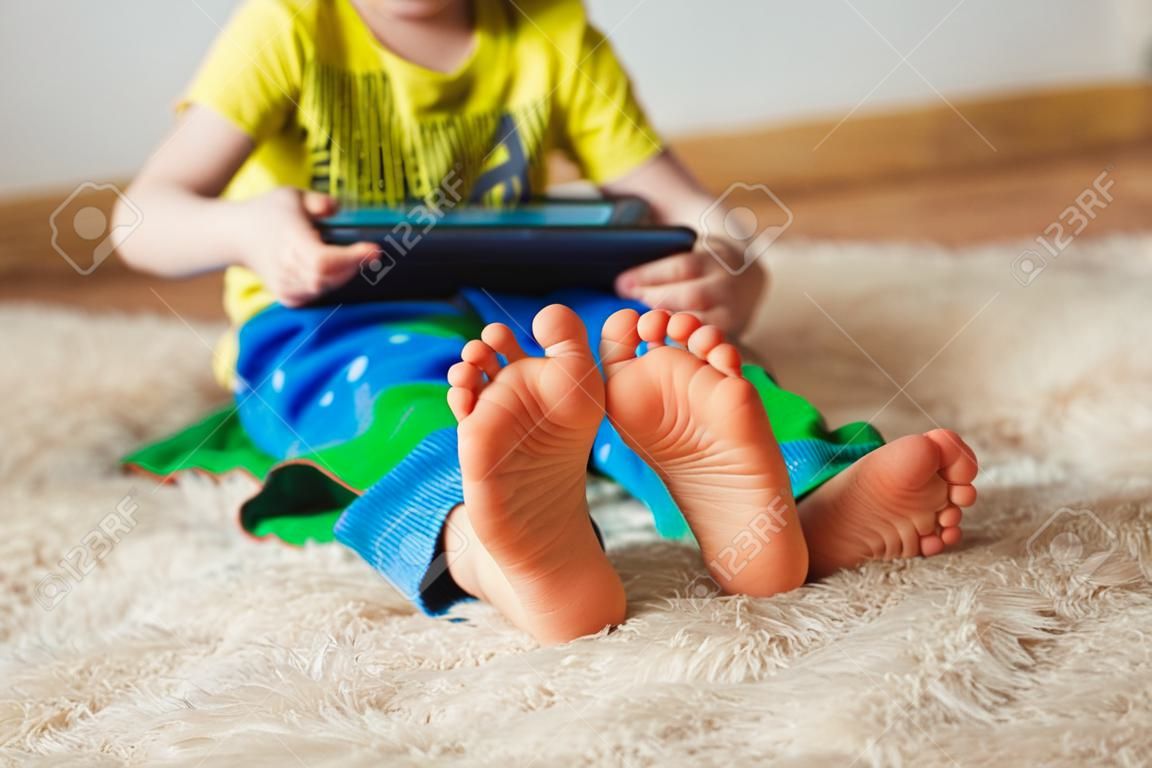Kind mit Tablette sitzen auf dem Bett und beobachten Karikaturen . Selektiver Fokus auf den Füßen . Nettes Junge im grünen T-Shirt und in der blauen Hose