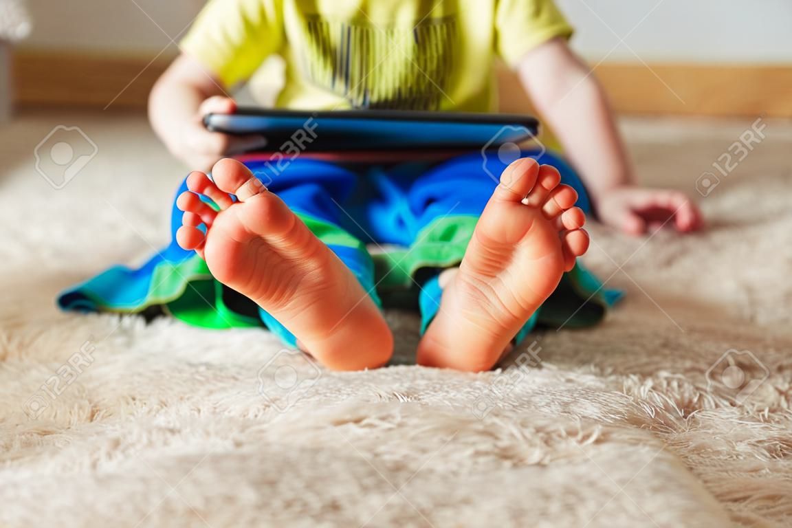 Kind mit Tablette sitzen auf dem Bett und beobachten Karikaturen . Selektiver Fokus auf den Füßen . Nettes Junge im grünen T-Shirt und in der blauen Hose