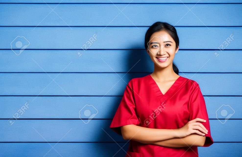 Portret przyjaznej, wesołej, uśmiechniętej, pewnej siebie azjatyckiej lekarki lub pielęgniarki w niebieskim garniturze stoi z rękami skrzyżowanymi na tle drewnianej ściany, pracownik służby zdrowia w niebieskich zaroślach z miejsca na kopię.