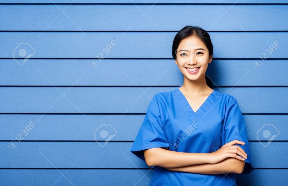 친절하고 쾌활하며 웃고 있는 자신감 있는 아시아 여성 의사나 간호사의 초상화는 나무 벽 배경에 팔짱을 끼고 서 있고, 복사 공간이 있는 파란색 수술복을 입은 의료 전문가입니다.