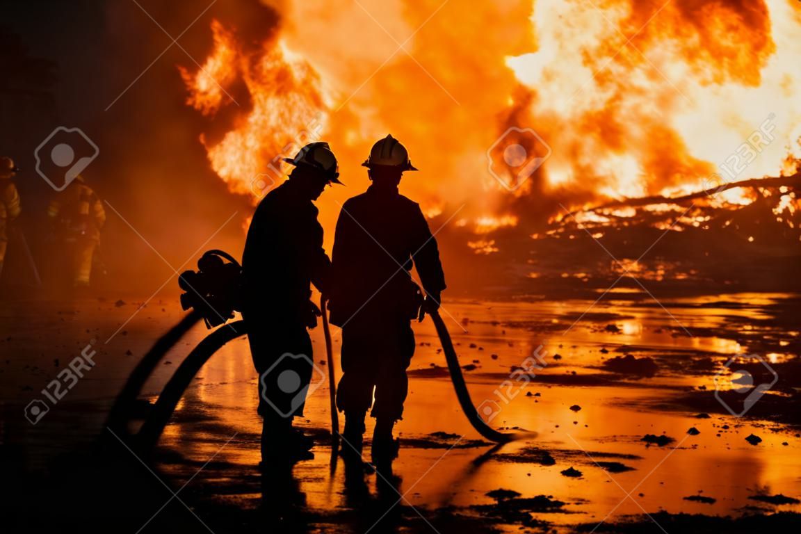 Schattenbild von den Feuerwehrmännern, die ein rasendes Feuer mit enormen Flammen des brennenden Bauholzes kämpfen
