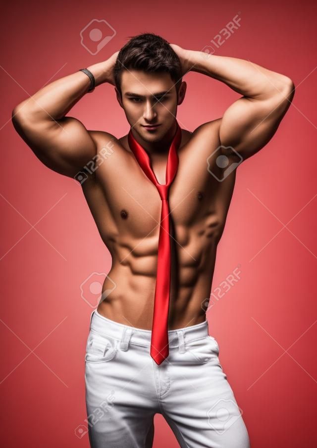 Atractivo joven hombre descamisado muscular, que llevaba sólo pantalones y corbata roja, aislado en blanco