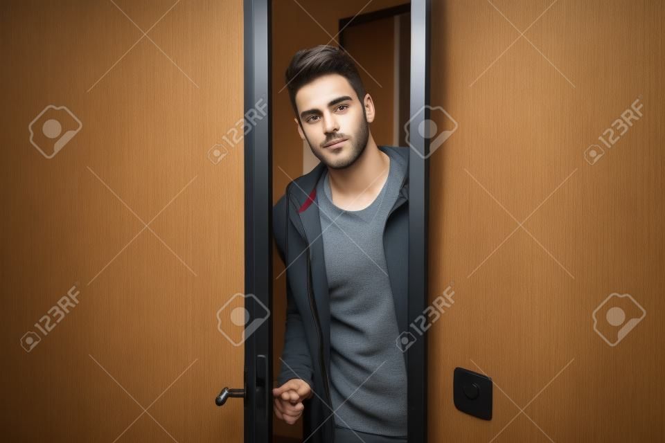 Jovem bonito abrindo a porta para entrar em uma sala, olhando para a câmera