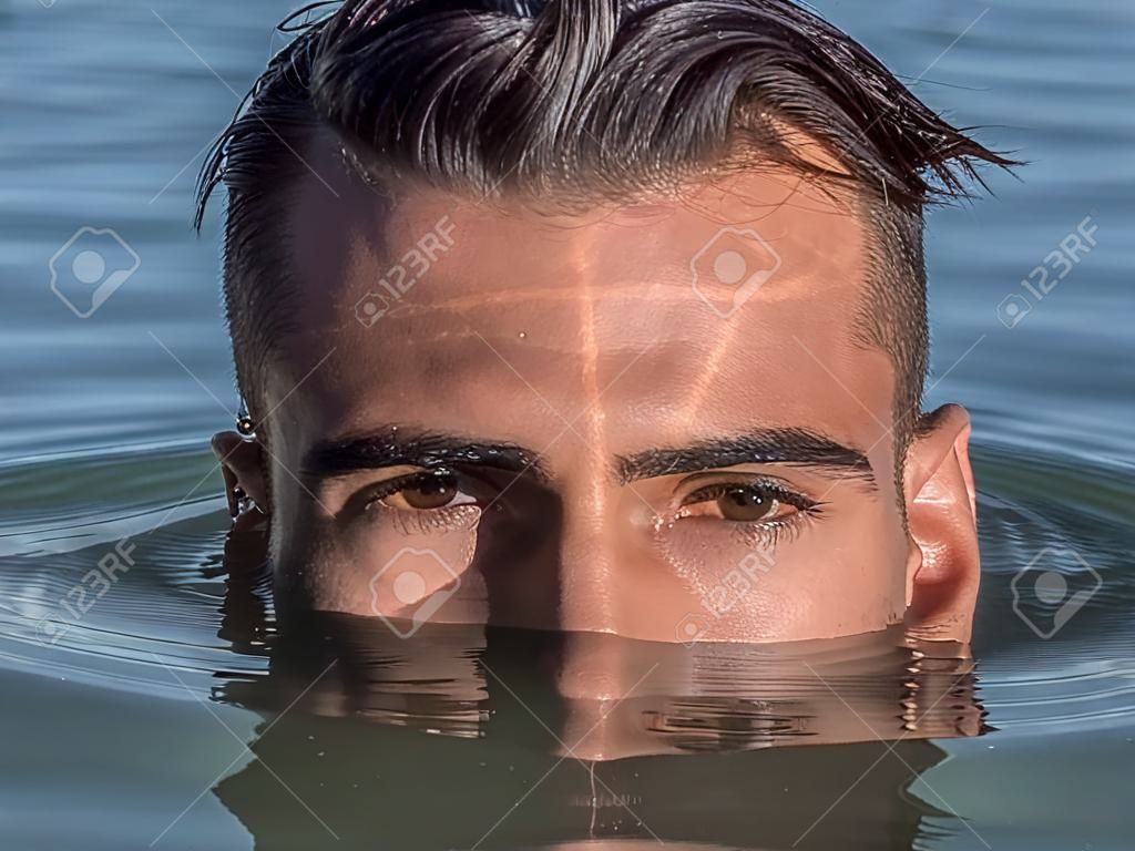 Attractive young shirtless athletic man standing in water in sea or lake, with half face submerged underwater, looking at camera