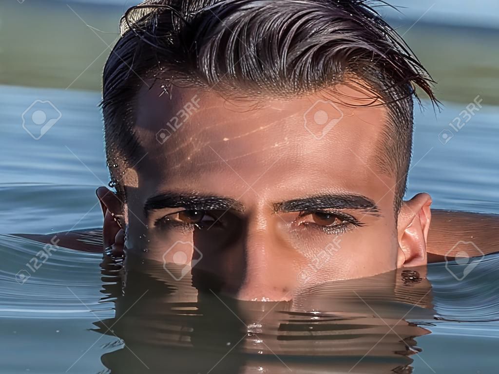 Attractive young shirtless athletic man standing in water in sea or lake, with half face submerged underwater, looking at camera