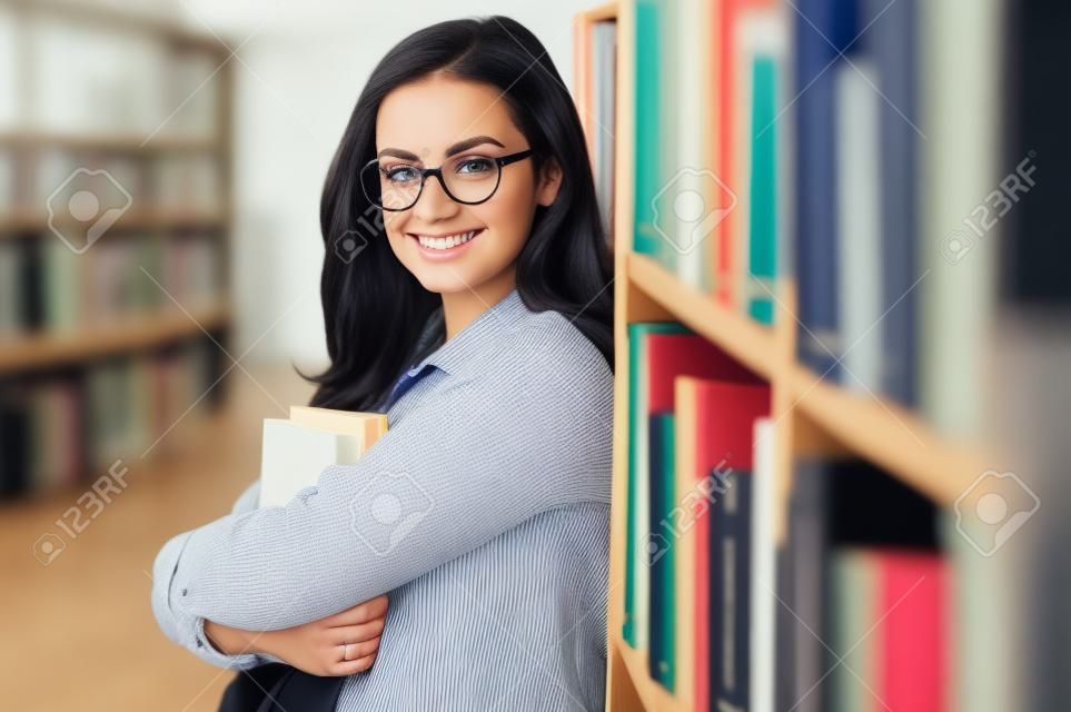 Portrait of young business female or student in eyeglasses, holding in arms some books, standing in library. Stylish caucasian female teacher looking at the camera, smiling, education concept