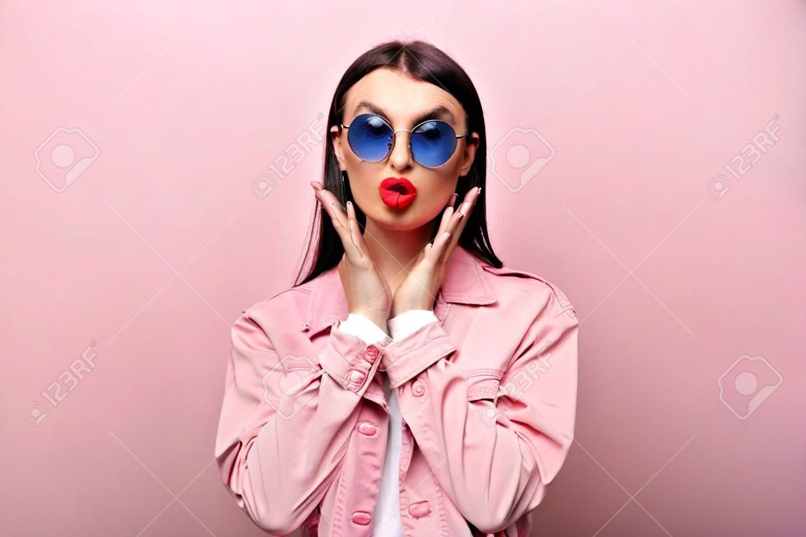 Atraente feliz elegante caucasiano jovem mulher em óculos de sol, posando no fundo rosa pastel isolado, mostra gesto de beijo
