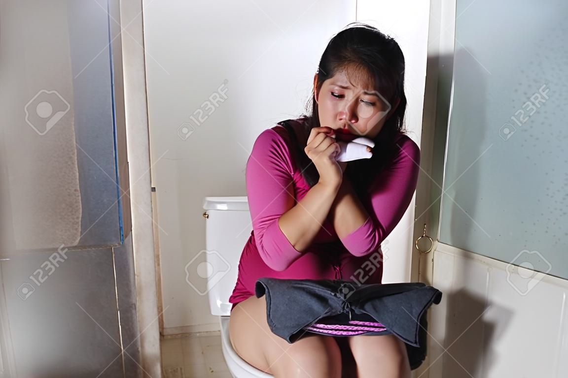 young scared and desperate pregnant Asian Korean woman or teenager checking positive result on predictor test sitting on toilet WC crying overwhelmed in unwanted pregnancy 