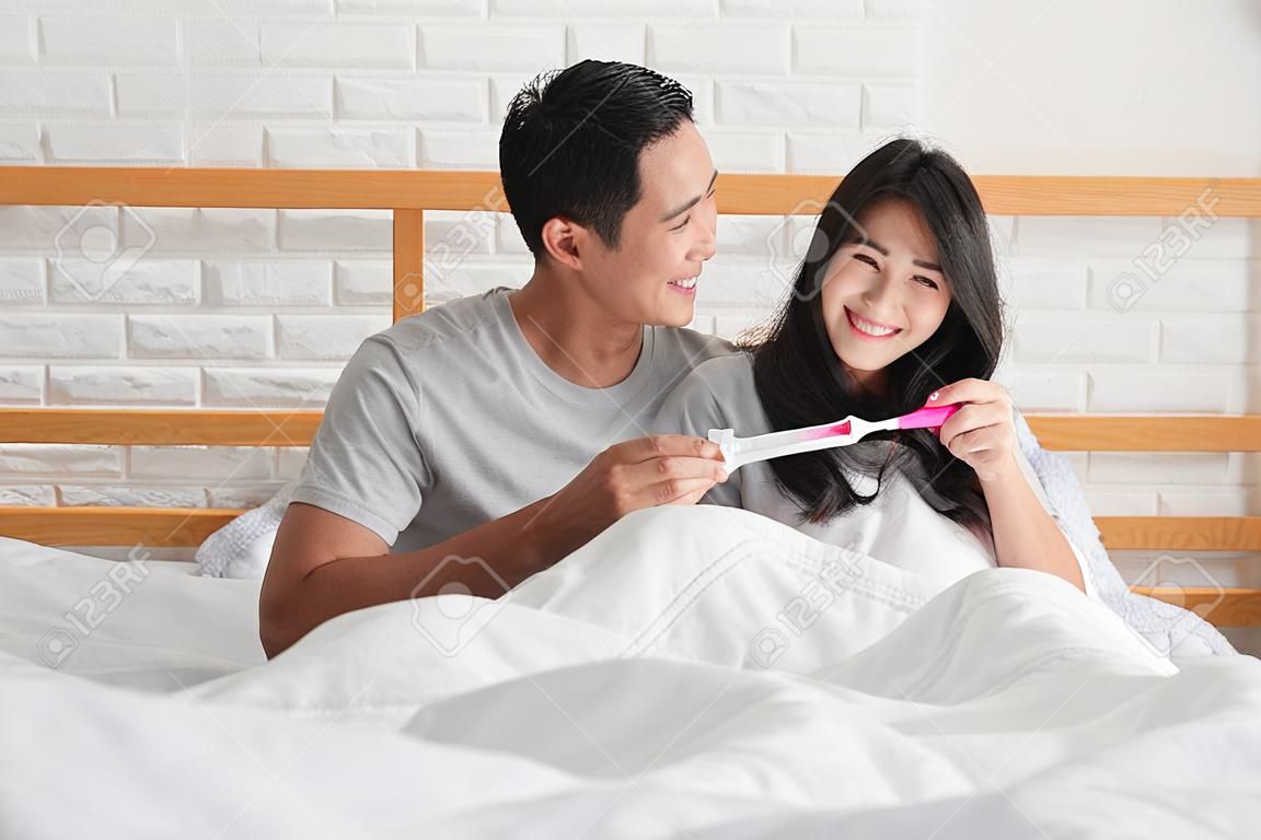 Casal asiático feliz sorrindo depois de descobrir teste de gravidez positivo no quarto em casa
