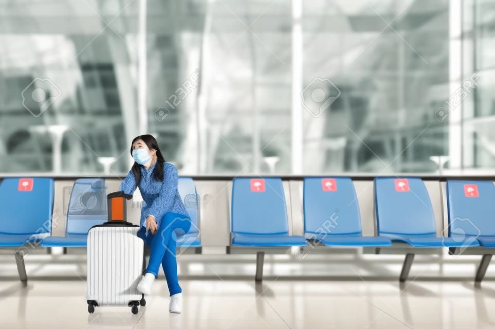 Asiatische touristin mit gesichtsmaske sitzt auf einem sozial distanzierenden stuhl mit gepäck und wartet auf den flug am flughafenterminal während des ausbruchs von coronavirus oder covid-19. Neue normale Reise am Flughafen
