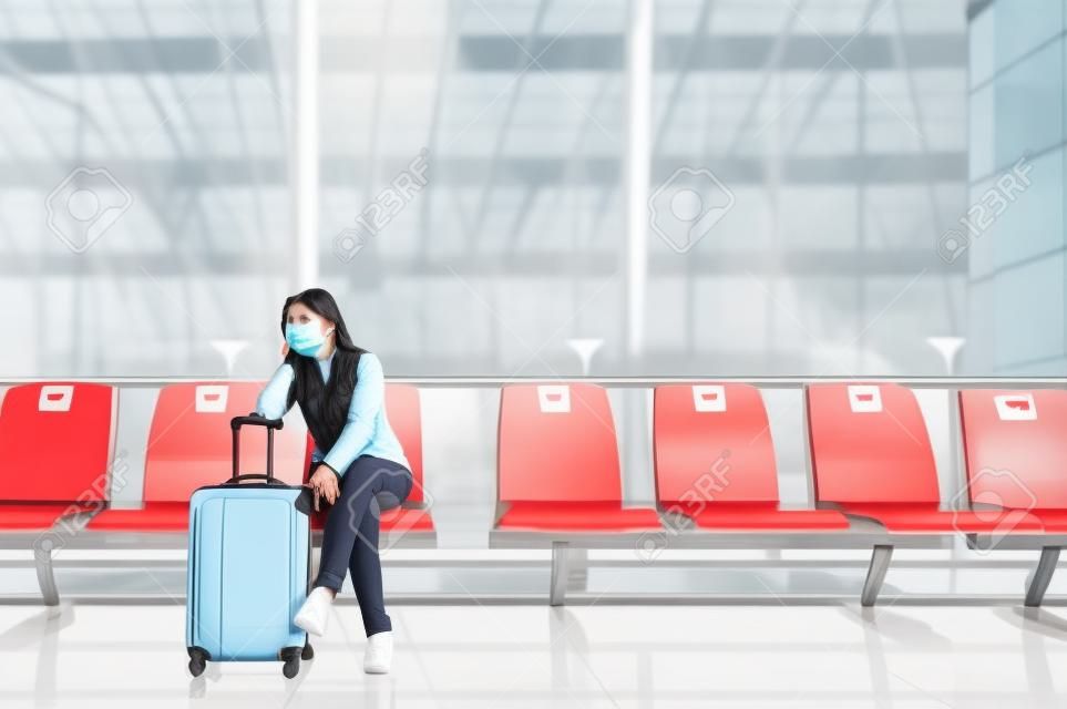 Asian woman tourist wearing face mask sitting on social distancing chair with luggage waiting for flight at airport terminal during coronavirus or covid-19 outbreak . New normal travel at airport