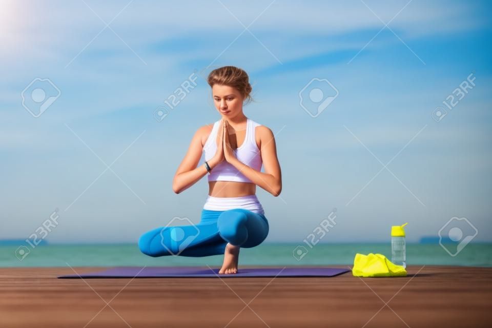 Sportliches junges Mädchen morgens auf dem Pier am Meer, Yoga praktizierend. Frau macht Gymnastik im Freien. Gesundheits- und Yoga-Konzept. Weibliche Meditation für Fitness-Lifestyle der Naturhintergrund