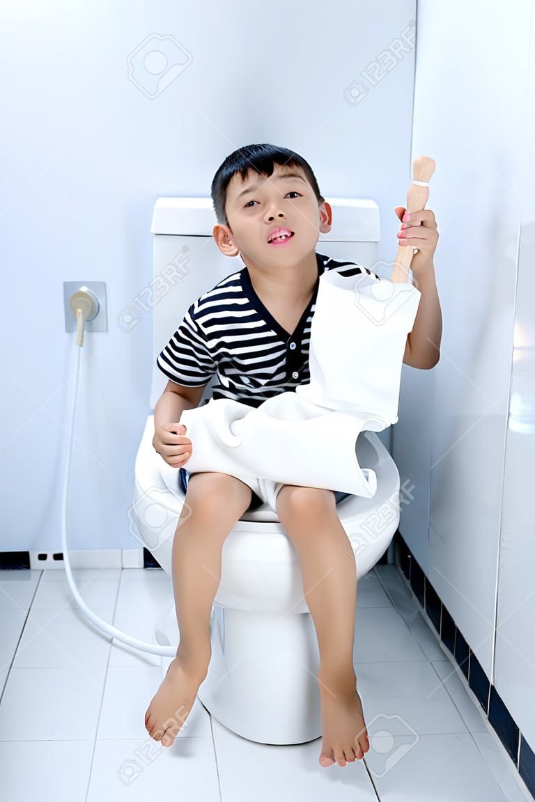 若いアジアの少年がトイレで白い洗面所の上に座る