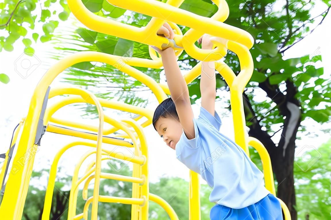 El muchacho asiático joven colgar la barra amarilla por su mano para ejercer en el patio puerta a cabo bajo el gran árbol.