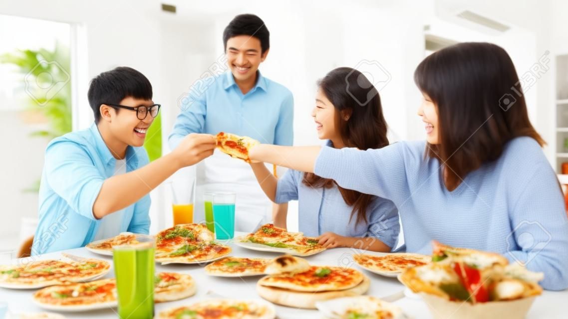 Fröhliche Gruppe junger Freunde, die zu Hause zu Mittag essen. Asiatische Familienfeier, die Pizzaessen isst und lacht und das Essen genießt, während sie zusammen am Esstisch im Haus sitzen. Feiertag und Zusammengehörigkeit.