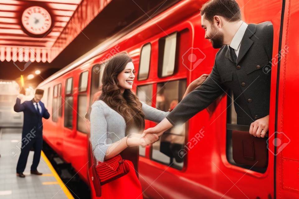 Coppia vintage, uomo in uniforme, donna in abito rosso, che si tiene per mano addio alla stazione ferroviaria mentre il treno parte