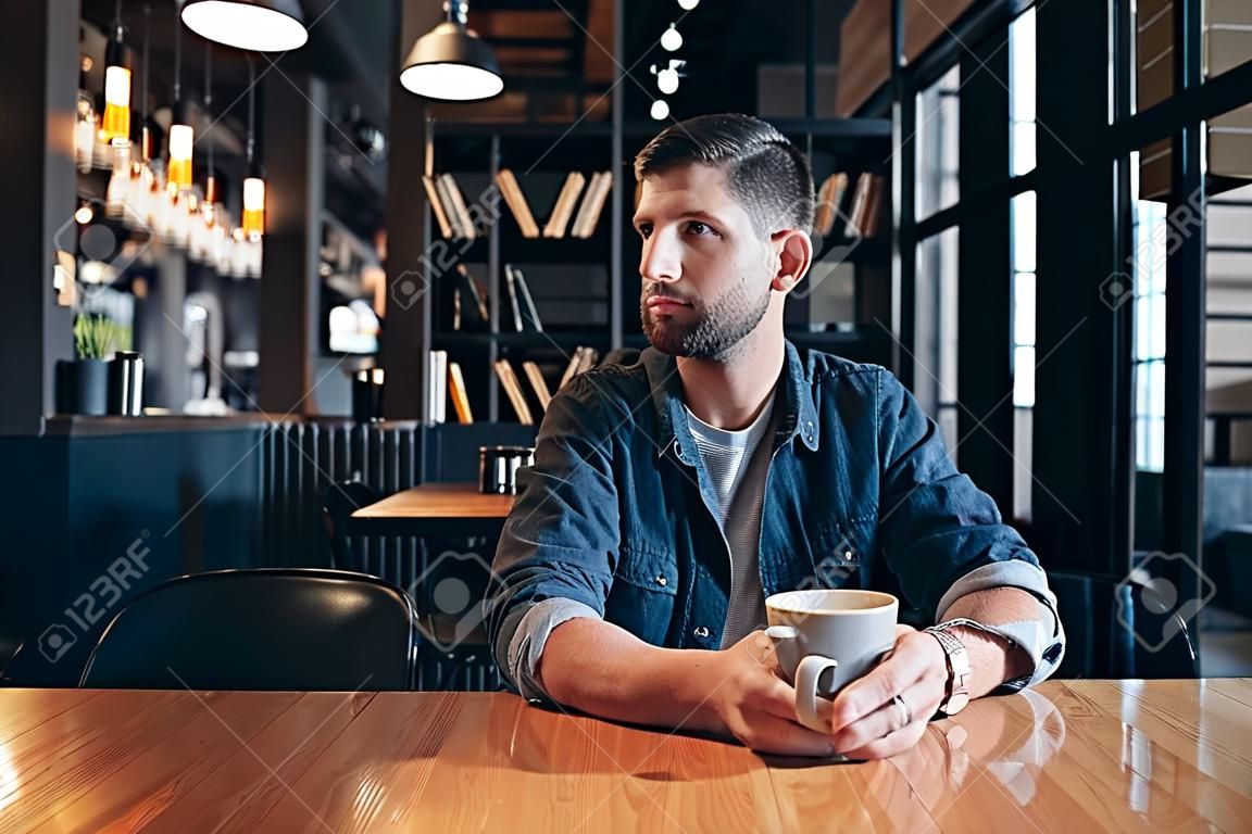 Selbstbewusster Mann, der eine Tasse Kaffee genießt, während er im Café im Innenbereich zu Mittag isst und nachdenklich aussieht