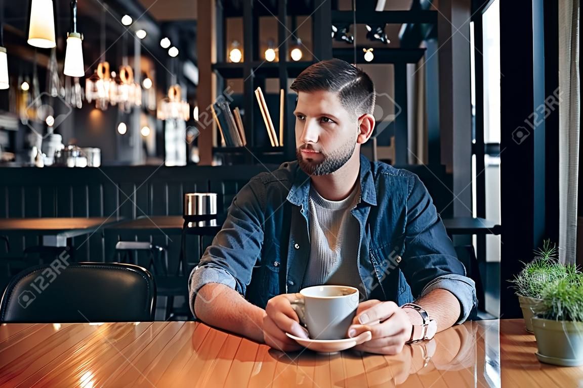 Selbstbewusster Mann, der eine Tasse Kaffee genießt, während er im Café im Innenbereich zu Mittag isst und nachdenklich aussieht