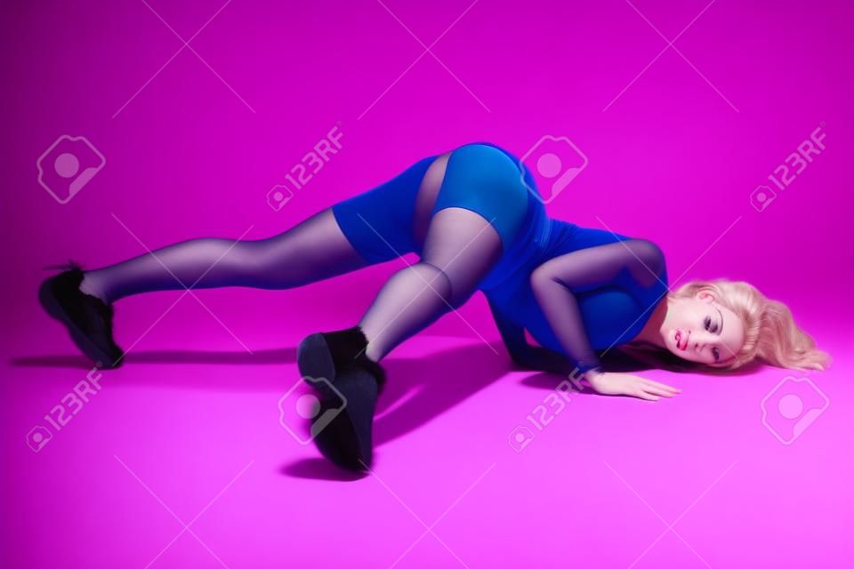 piękna kaukaska blondynka w welurowych niebieskich spodenkach i czarnych pończochach tańczy twerk leżąc na różowym tle