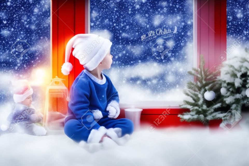 Criança bonito, sentado em uma janela, olhando ao ar livre para o Papai Noel, decoração de Natal na janela