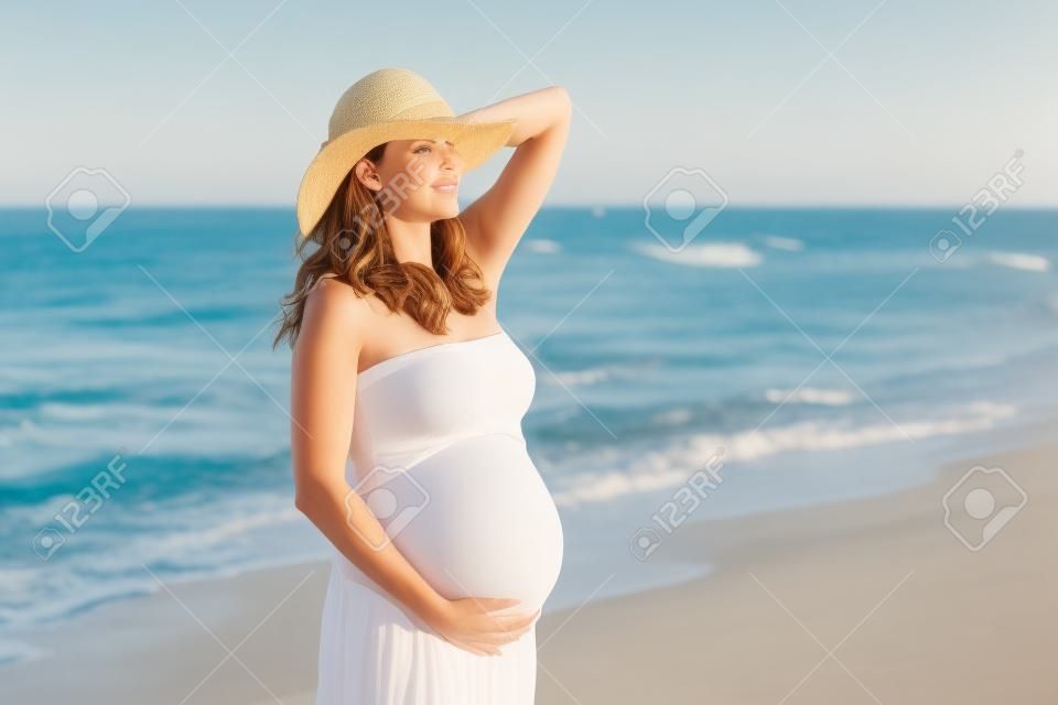 Beautiful pregnant woman on the beach with hat touching her belly