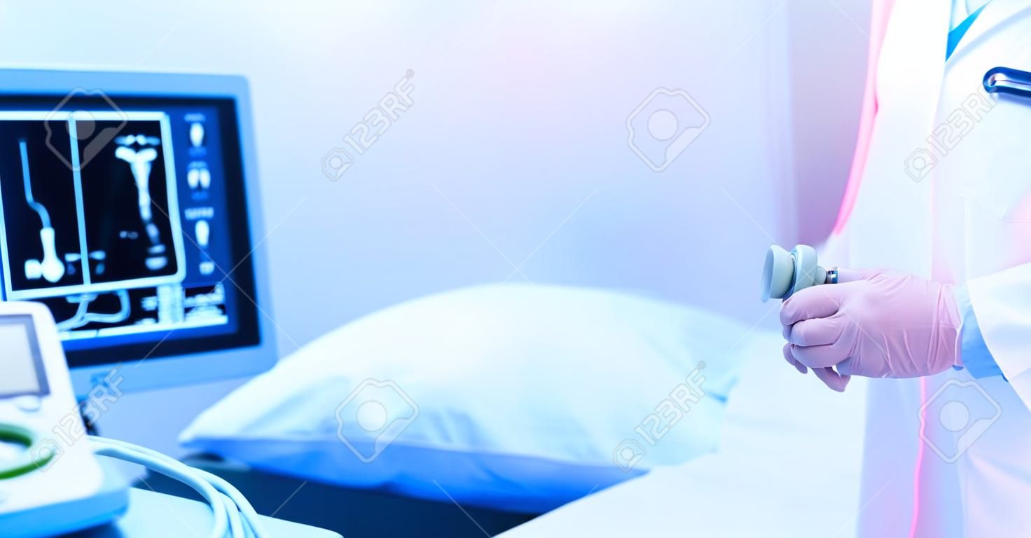 Primo piano medico che tiene uno stetoscopio nella foto in tonalità blu della macchina ad ultrasuoni che viene utilizzata per l'esame diagnostico del paziente. Messa a fuoco selettiva sulla sonda con sfondo sfocato. Sfondo di concetto medico