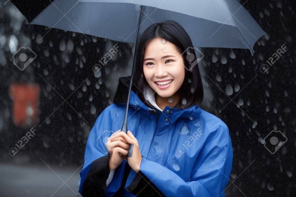 Asiatische Frau des regnerischen Tages, die draußen einen Regenmantel trägt. Sie ist glücklich.